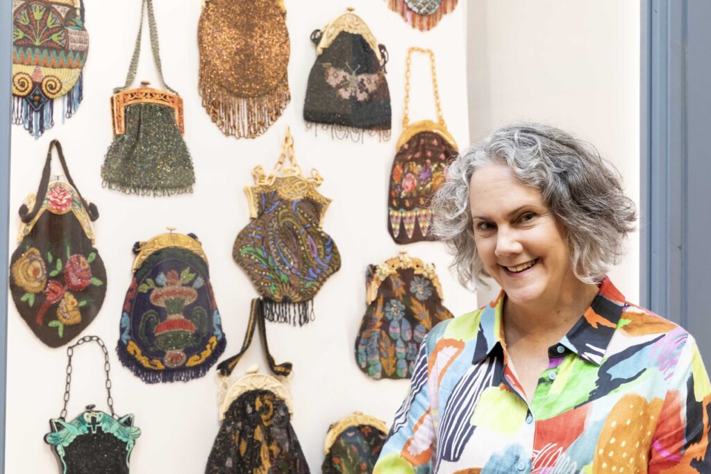 A woman stands in front of an array of antique beaded purses hanging on a white museum wall.