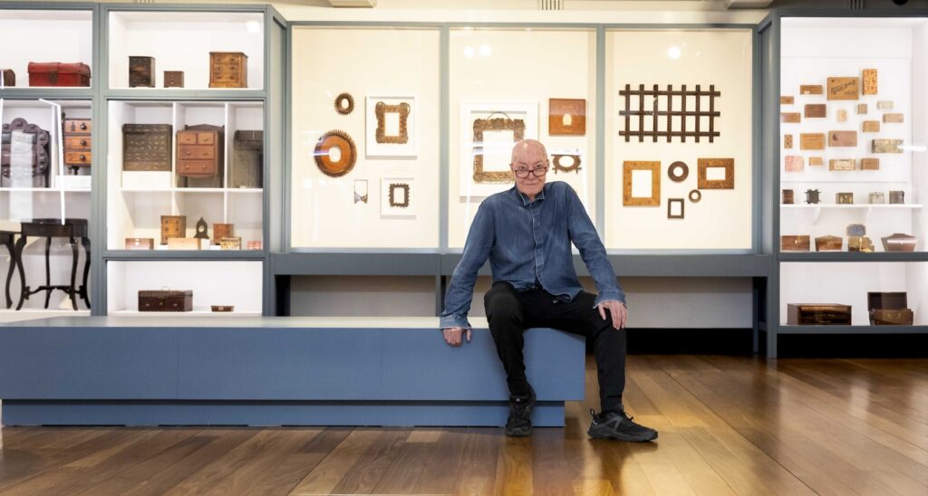 A man sits on a low bench in a museum. Behind him glass cabinets are filled with various wooden objects and furniture.