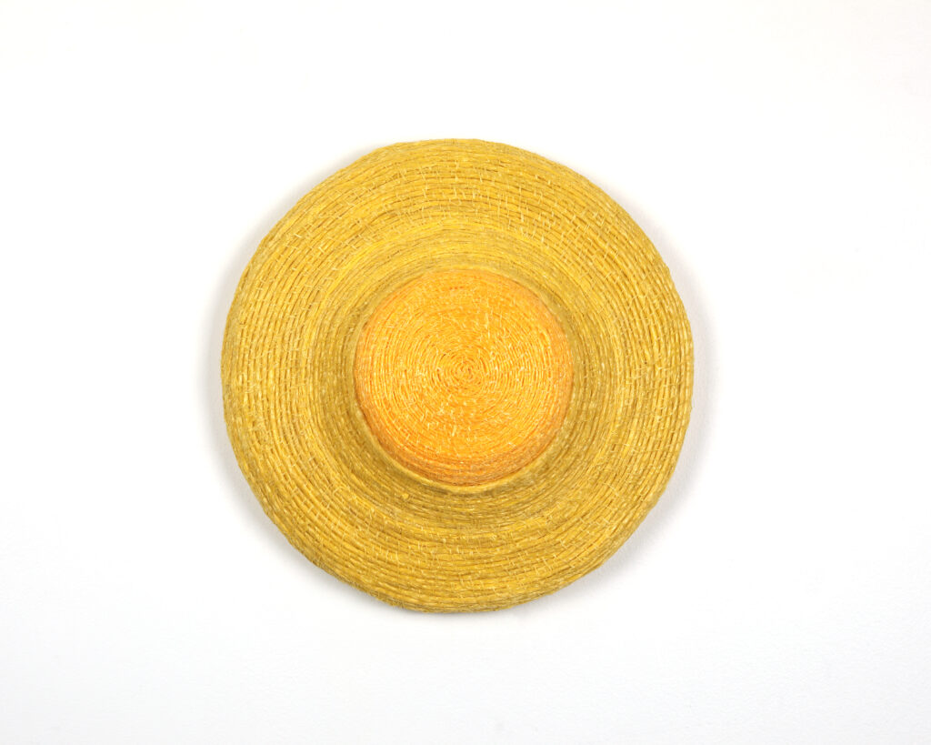 A bright, yellow disc woven from baling twine upon a white background. The disc has a lighter yellow on the outer perimeter and a darker yellow in the centre.