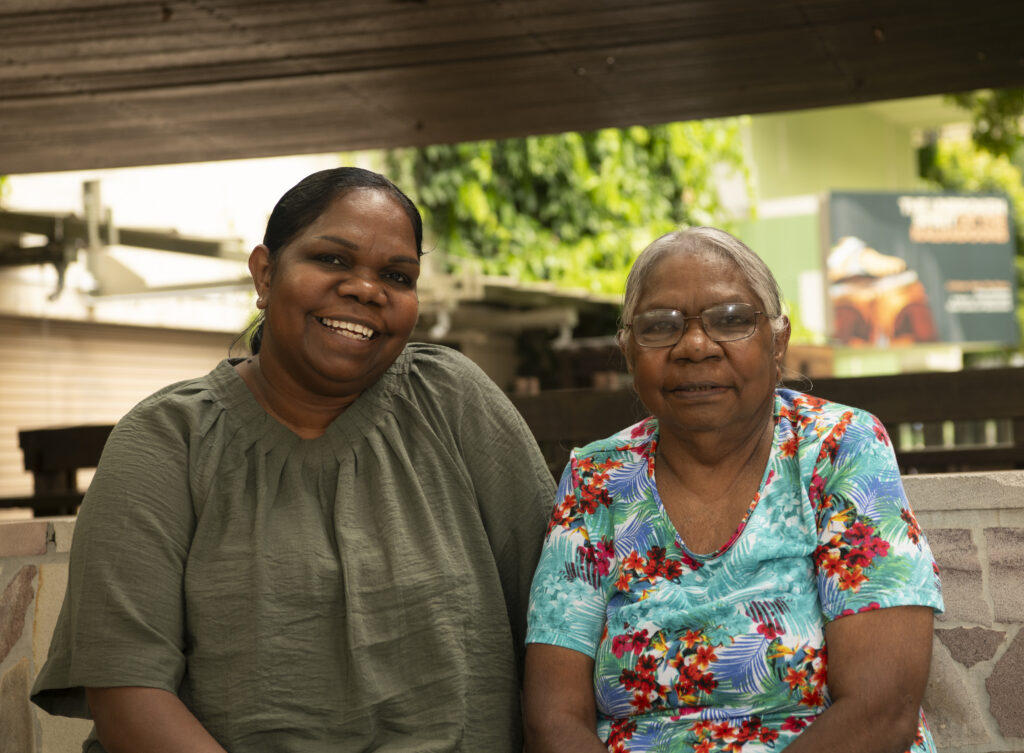 A photograph of Colleen Sam and Aunty Ena. Sam is seated on the left wearing an olive green blouse. Aunty Ena is seated on the right wearing a bright teal blouse with a floral design.