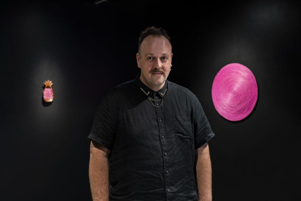 The artist stands before two of his works in the gallery. To his right is a bright pink disc; to his left is a smaller pineapple sculpture in tones of bronze and pink. The artist is wearing a black collared shirt with silver, star-shaped embellishments on the collar.