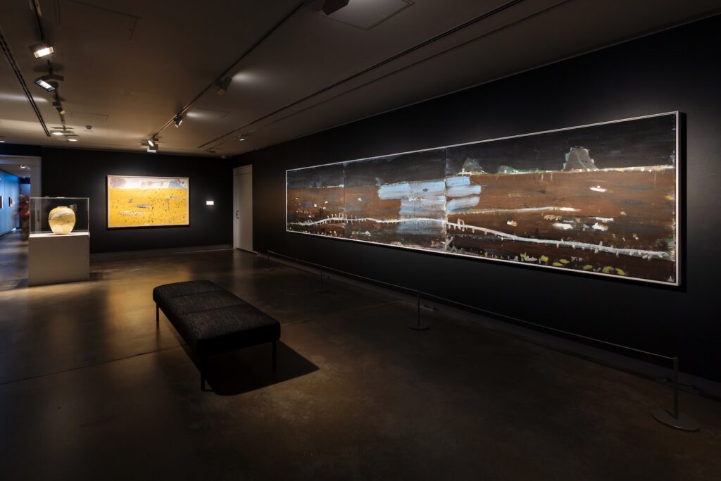 A gallery space with dark black walls and warm ambient spotlights on three artworks. Two landscape paintings hang on separate walls and one white clay ceramic vase sits on a plinth in a clear glass case. In the centre of the room is a low, black fabric bench.