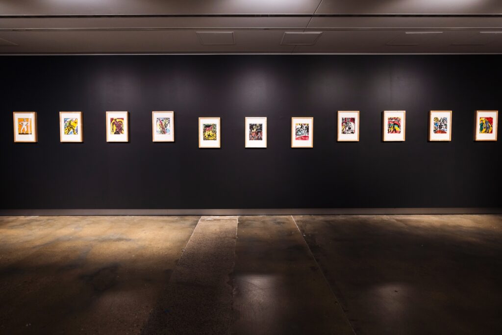 A gallery space with a shadowed concrete floor. Hanging on a dark coloured wall, warm ambient spotlights illuminate a single row of eleven, small, multicoloured woodcut artworks depicting different circus images. The circus acts from left to right are titled Laughing clown, Strong man, Russian gymnast, Balancing on a ball, Clown on a goat, Tumblers, Horses in the arena, Violist, Trapeze, Ring master, 2 clowns.