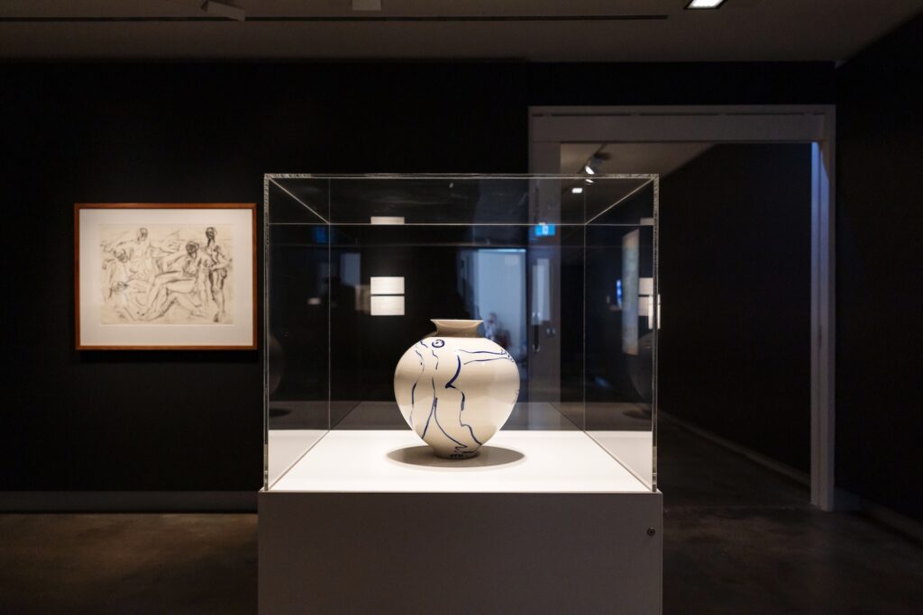 A gallery space with dark black walls and warm ambient spotlights on two artworks. In the foreground is a white ceramic vase with a human figure painted in cobalt blue. The vase sits on a white plinth under clear glass. In the background hangs a sketch of five human figures in various seated and standing positions.