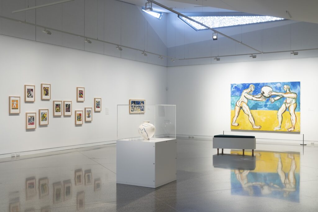 A light-filled gallery space with white walls and three artworks. In the centre of the room is a white vase on a plinth in a glass case. On the far wall is a large blue and yellow painting of two figures. On the left wall is a jumbled series of eleven, small, multicoloured, woodcut artworks depicting different circus images.