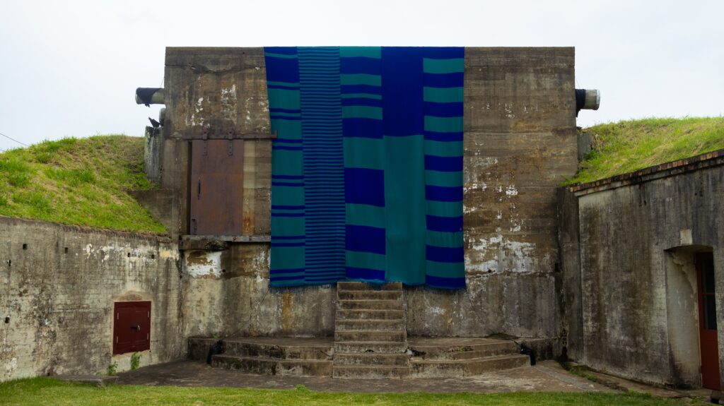 A large teal and navy blue textile hangs from a weathered concrete structure. The textile has a geometric pattern of stripes and blocks. The concrete structure is set within a grassy landscape.