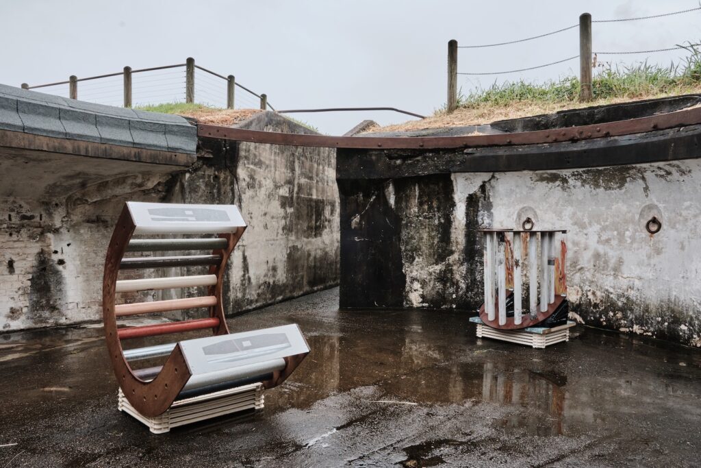 Two sculptural installations sit in an outdoor concrete area. The photo has been taken after rain and all the surfaces are wet. The first sculpture is a curved, wooden structure with multiple tubes, and translucent panels at the top and bottom. The second sculpture, smaller and more compact, is also made of two curved, wooden sides and connecting tubes.