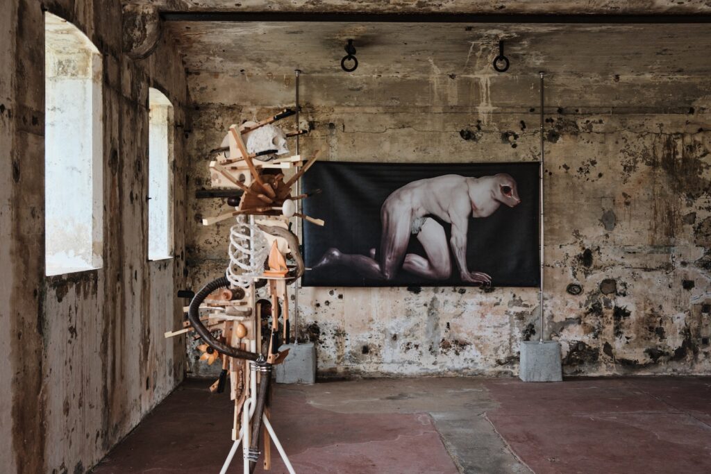 This image shows a large dilapidated room with concrete walls and a reddish-brown floor. On the centre left is a sculpture made of assembled objects, pieces of timber and parts of a skeleton. At the back of the room is a large-format print depicting a human figure with an animal-like head crouching on all fours.