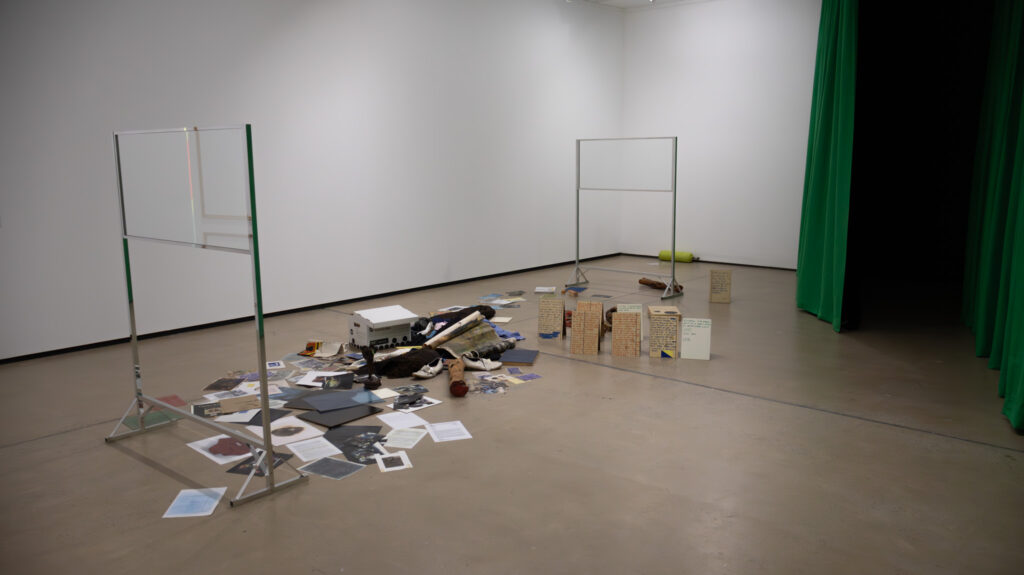 A pile of personal belongings is scattered chaotically across the gallery floor, bookended by glass screens on aluminium frames. A green curtain segments the perimeter of the gallery.
