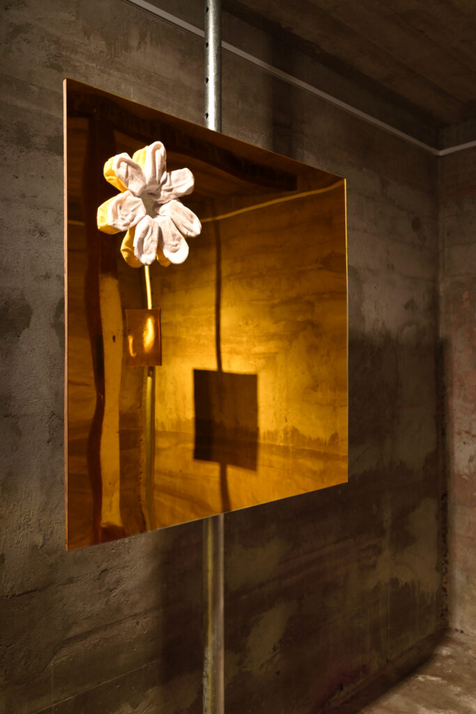 Large yellow rectangle reflecting golden light into the room around it. It is affixed in mid-air by being attached to one beam of grey metal scaffolding reaching floor to ceiling. The rectangle’s surface is made of yellow acrylic and in the top left corer a fist-sized hole has been cut out and in it arranged white ankle socks to give the appearance of a daisy.