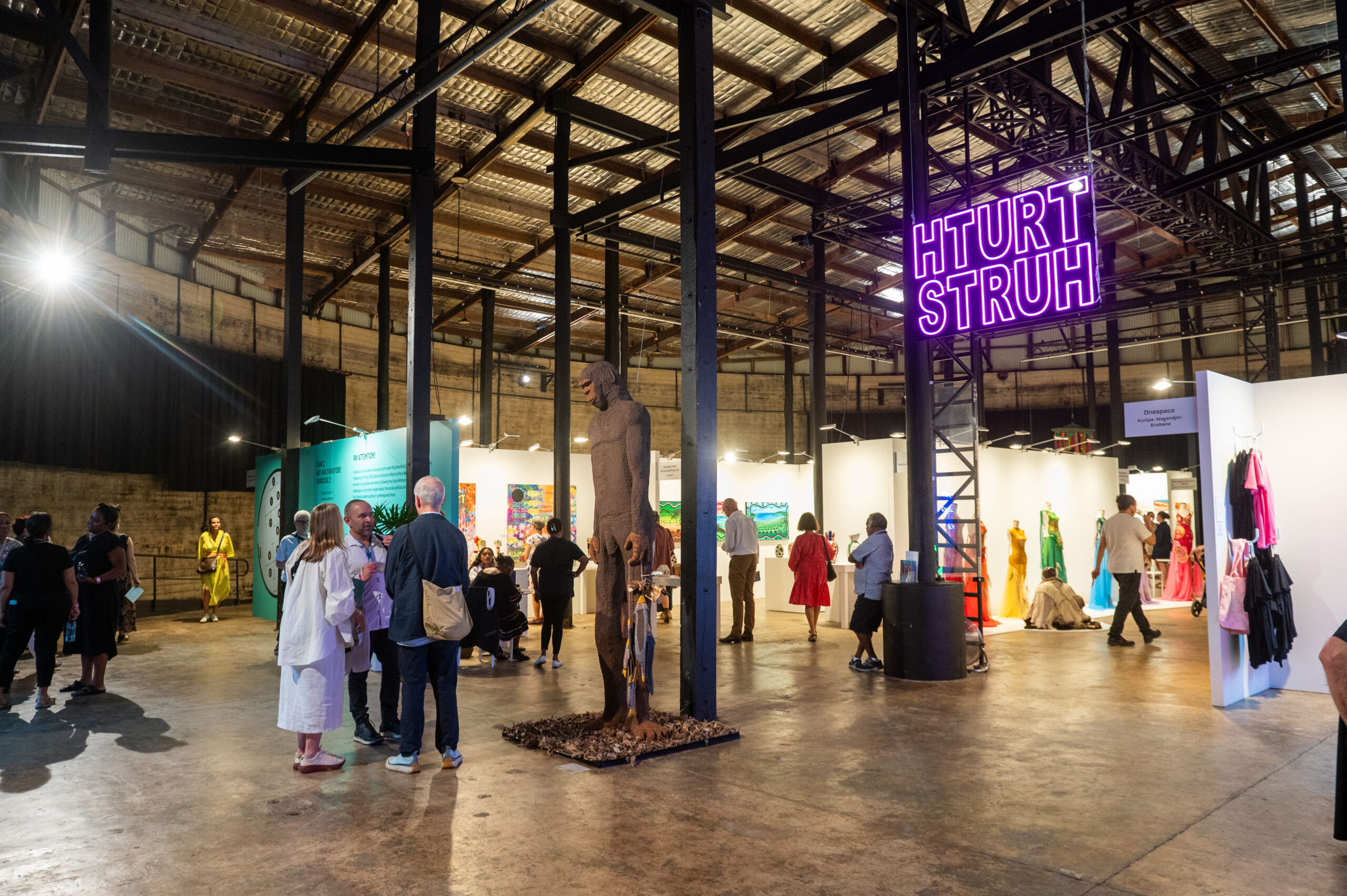A wide shot of a large industrial space full of artworks. People are gathering in front of works and talking to one another about them.
