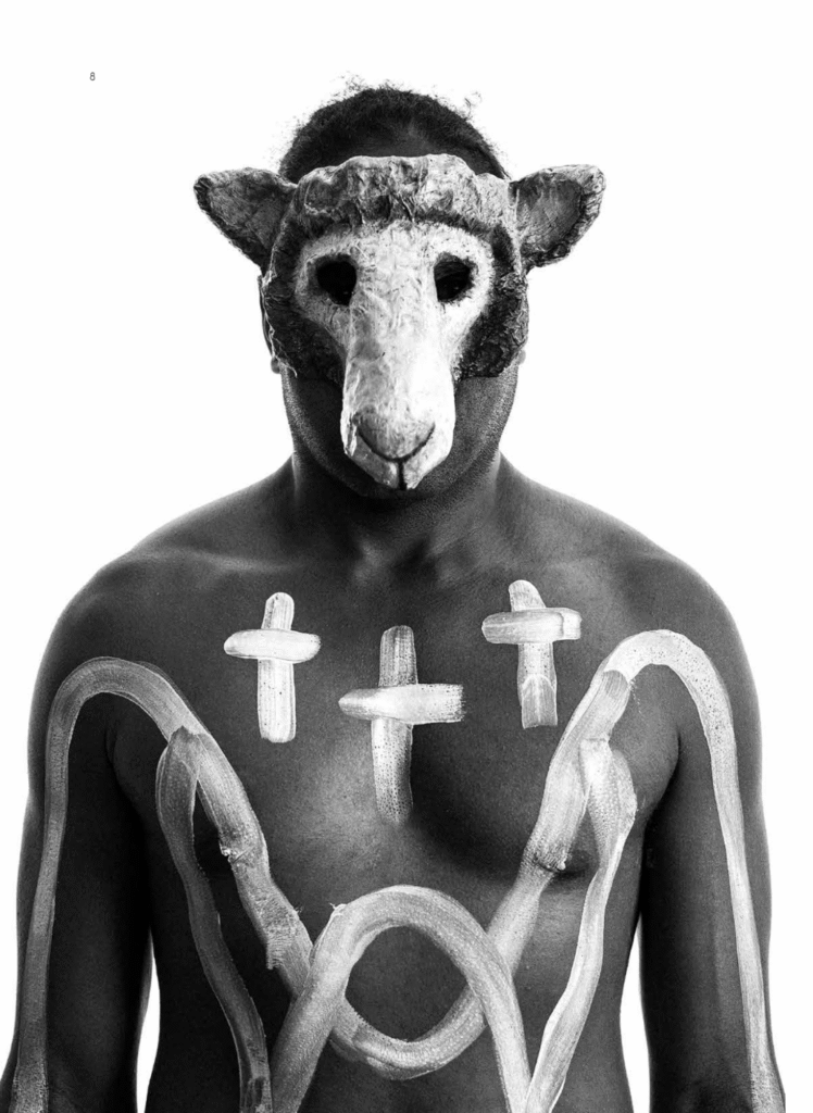A black and white photograph of an Aboriginal man wearing a sheep mask. He has three crucifixes painted on his chest.