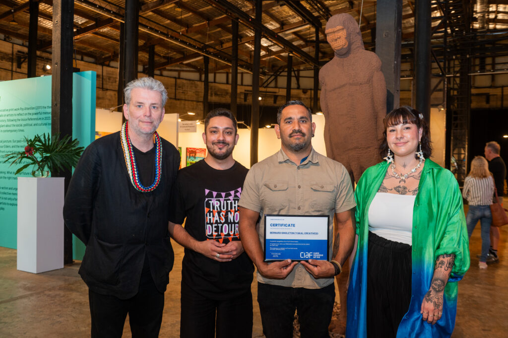 A group of four people stand before the artwork chosen for the Premier's Award for Excellence. The winner, Bernard Singleton, holds the winning certificate.