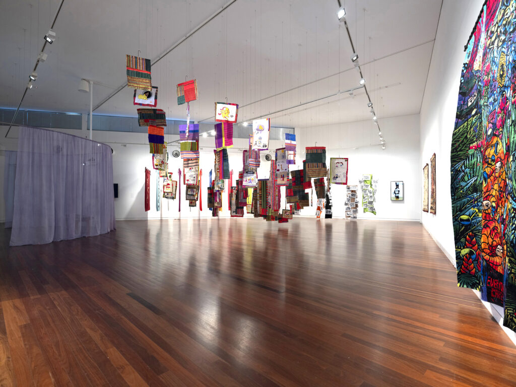 An installation view of Transfer. Textiles of various shapes and colours are displayed throughout a gallery space.