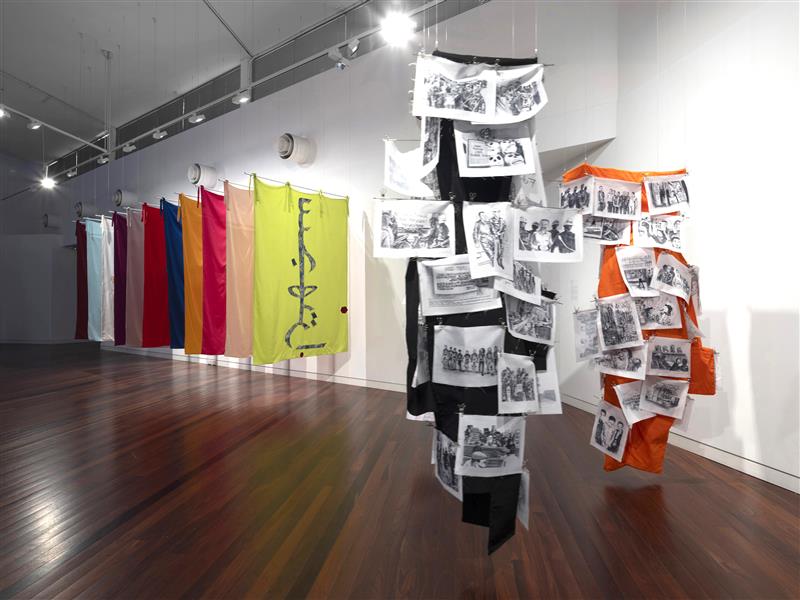 Columns of orange and black textiles are suspended from the ceiling. They are covered in textiles bearing illustrations. In the distance are a series of coloured textiles in a row.