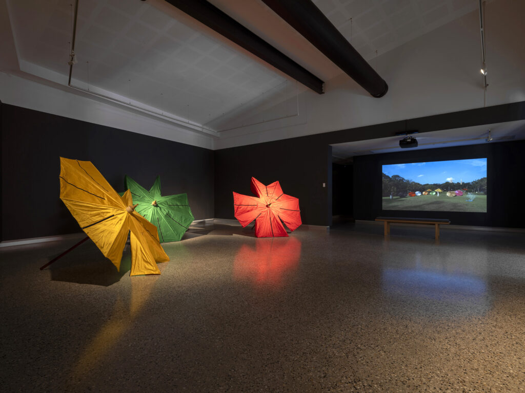 Three giant parasols lay on their sides in the gallery space. One is yellow, one is green, one is red. A film depicting parasols and other sculptural works is playing in the distance.