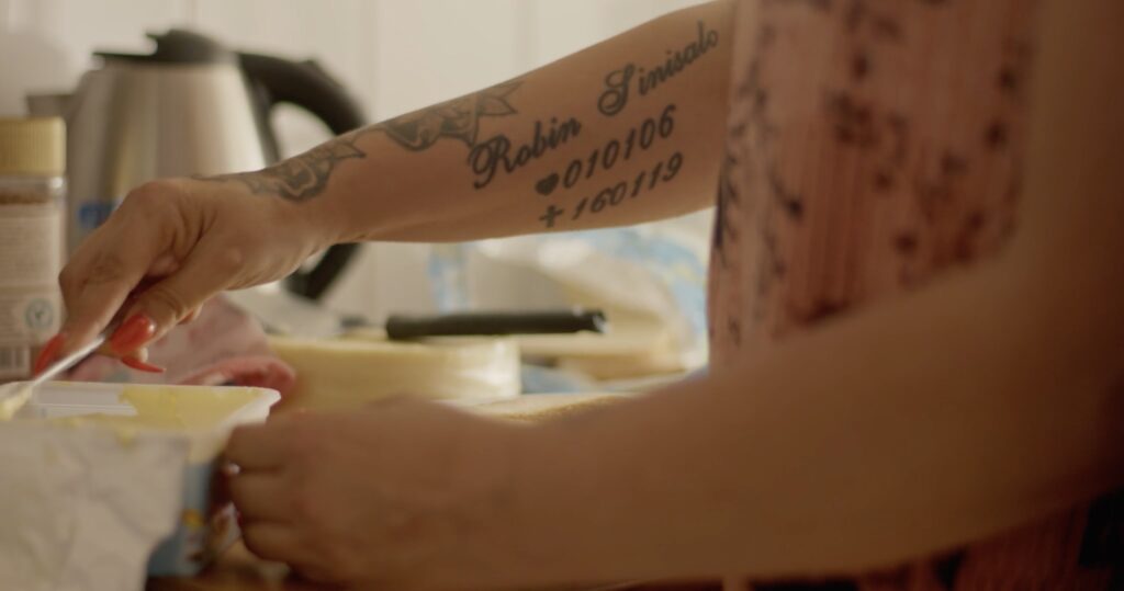 video still of a woman’s arm as she cuts a slice of butter in the kitchen, the focus on an arm tattoo in memoriam of her deceased child.