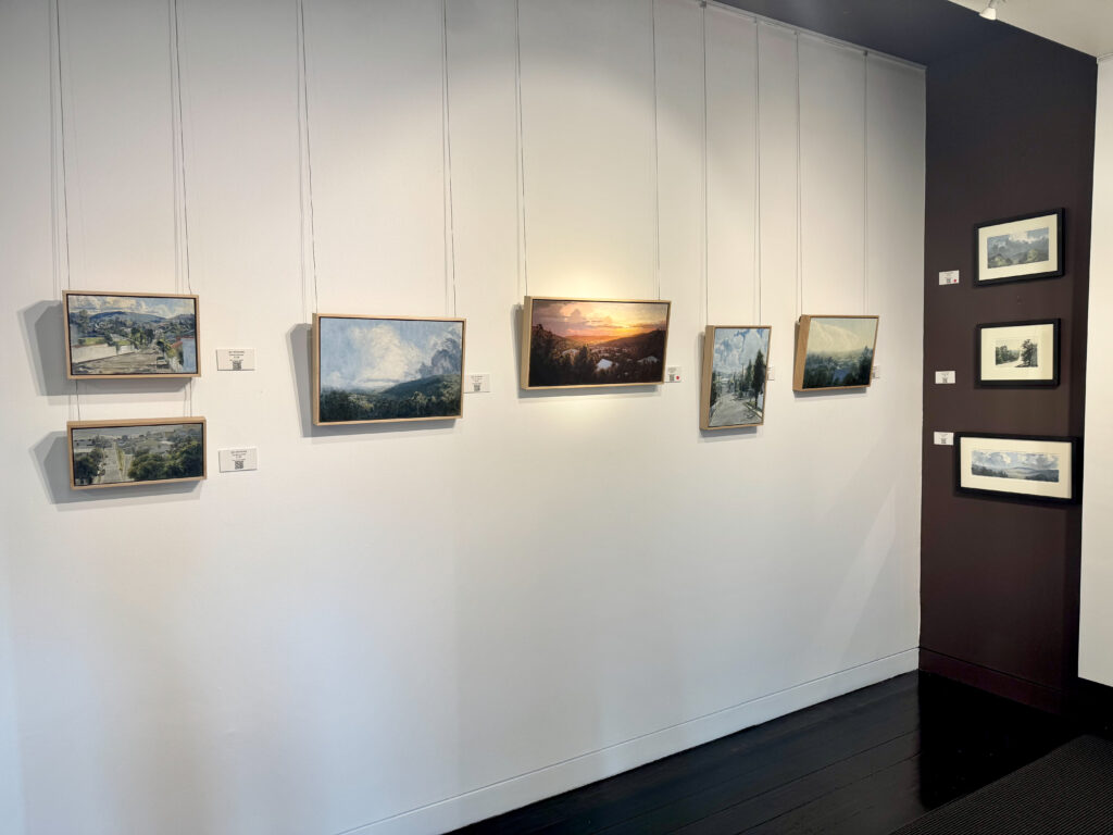 Nine small landscape paintings hung on two walls in a gallery